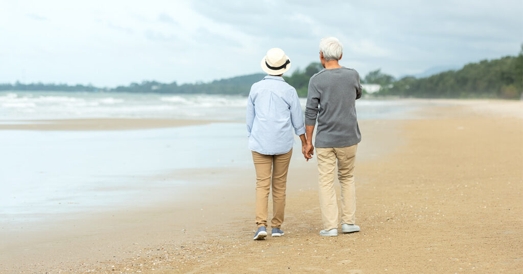 Retired couple enjoying a sunset stroll along the sandy beach, holding hands and smiling.