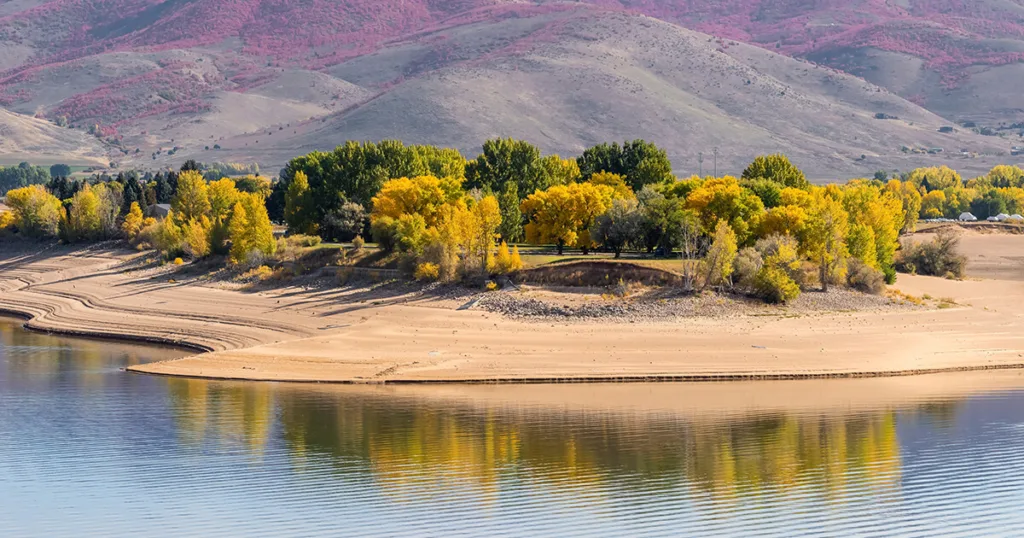 A breathtaking view of Pineview Reservoir in Ogden, Utah, surrounded by lush green trees. The crystal-clear blue water reflects the beautiful mountain landscape, offering a serene setting for camping. The Cottonwood Forest can be seen in the background, providing shady spots for tents and RVs.