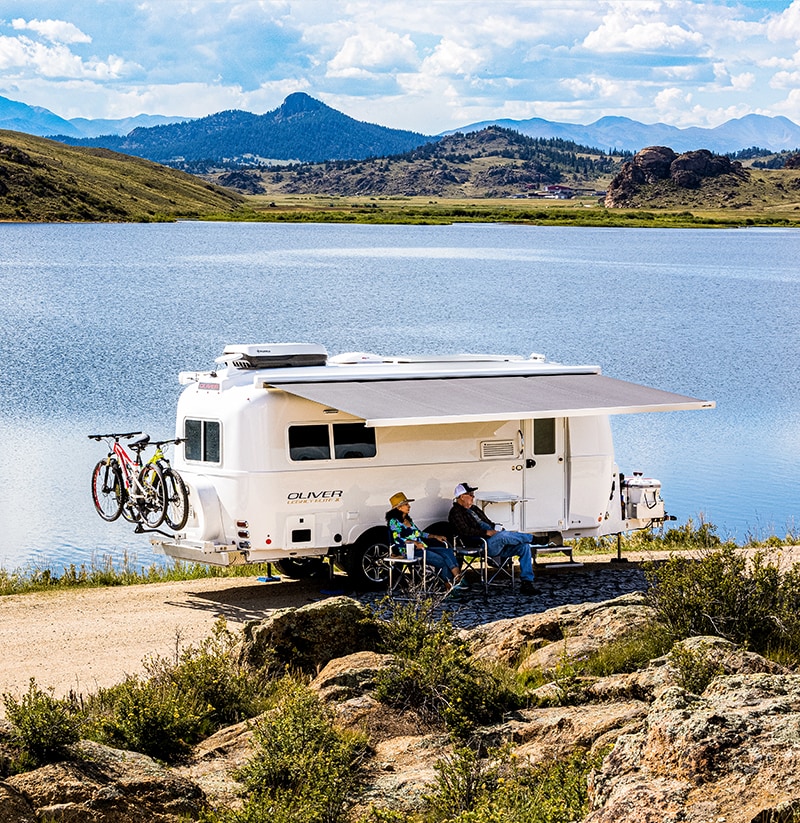 A couple sitting with their travel trailer near a lake on a sunny day.