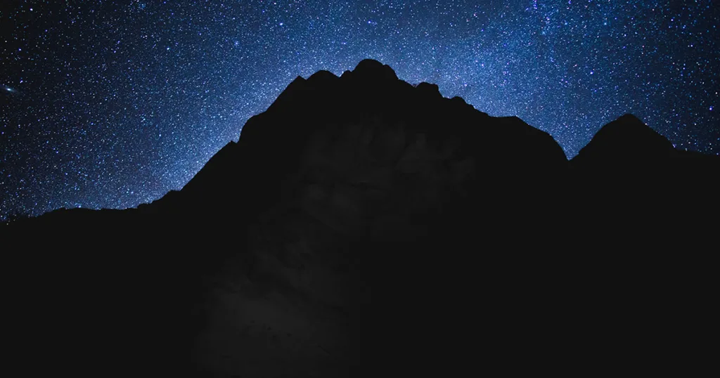 The clear night sky is adorned with countless twinkling stars, forming a mesmerizing celestial display. Silhouetted against the dark sky, the towering red rock cliffs of Zion National Park create a striking contrast. 