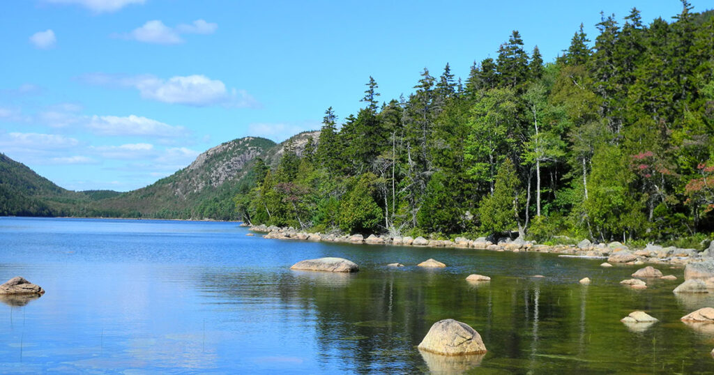 On a bright summer day, a picturesque view of Jordan Pond in Acadia National Park, Maine. The serene waters reflect the azure sky, fluffy clouds, and rugged rocks, creating a stunning natural landscape